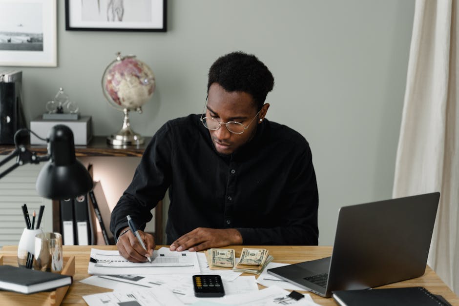 An adult man calculates expenses, using a laptop and documents at a desk in a home office setting.